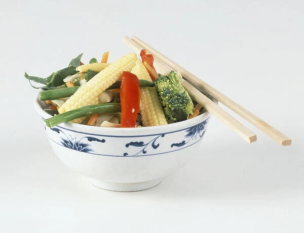 Bowl of vegetables, baby corn, peppers, green beans, carrots, broccoli, chopsticks resting on the rim of the bowl