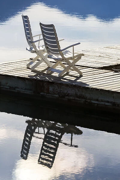 Two White Wooden Deck Chairs On Wooden Boat Dock Reflecting In The Water; Invermere, British Columbia, Canada