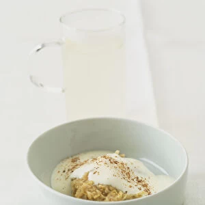 Boiled porridge oats served in white ceramic bowl with live natural yoghurt, sprinkled freshly grated nutmeg, accompanied by glass of hot water with added lemon juice