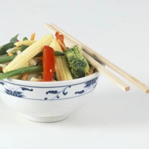 Bowl of vegetables, baby corn, peppers, green beans, carrots, broccoli, chopsticks resting on the rim of the bowl