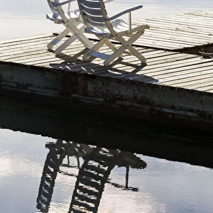 Two White Wooden Deck Chairs On Wooden Boat Dock Reflecting In The Water; Invermere, British Columbia, Canada